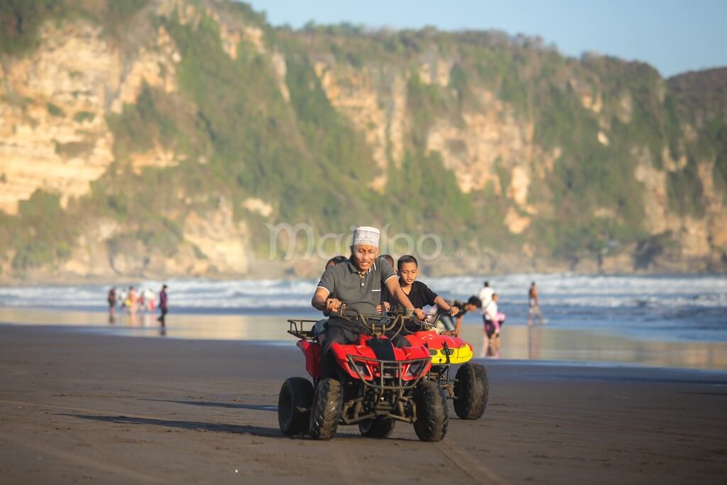 Two people riding ATVs on Parangtritis Beach with cliffs in the background.
