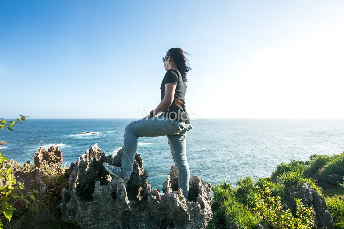 A tourist standing on a rocky cliff overlooking the ocean at Nglambor Beach, enjoying the view.