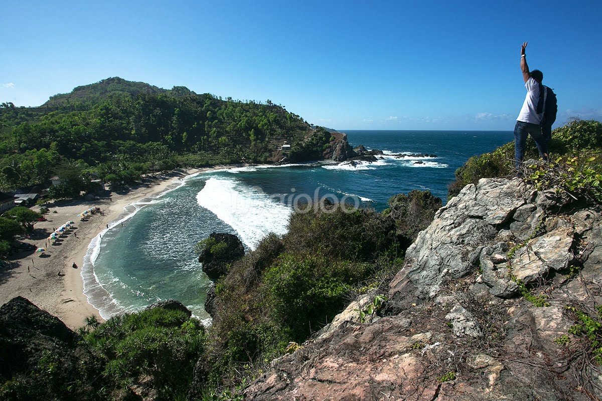 A person stands triumphantly on a cliff overlooking Siung Beach, with the ocean and lush green hills in the background.