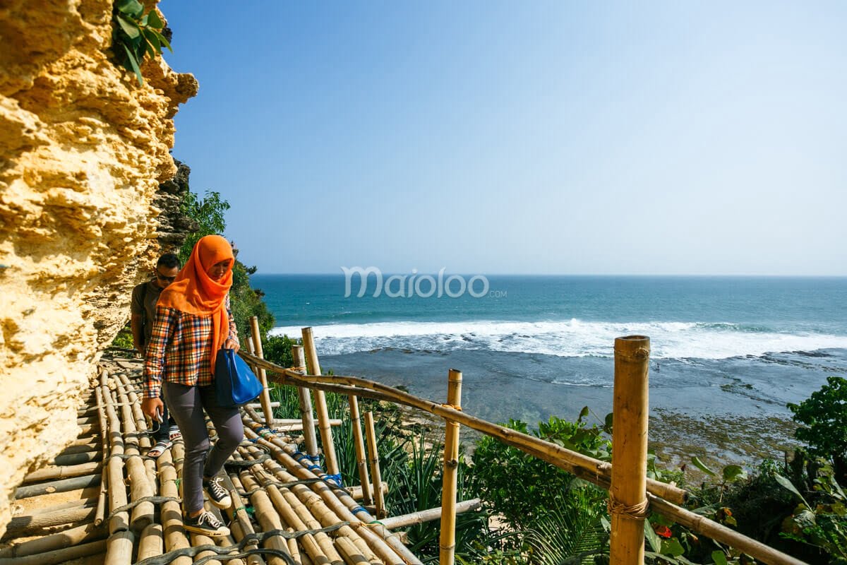 A woman and a man walking on a bamboo path along the edge of a cliff at Pok Tunggal Beach.