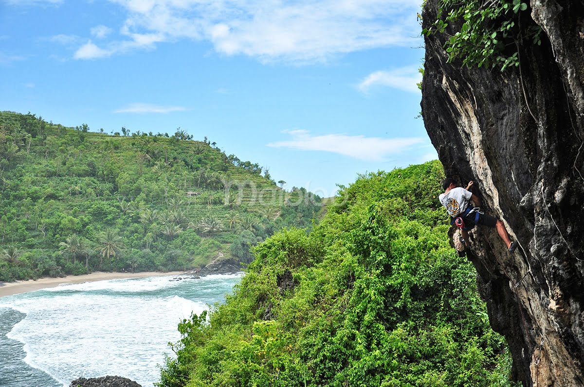 A climber scales a steep rock wall at Siung Beach with the ocean and green hills in the background.