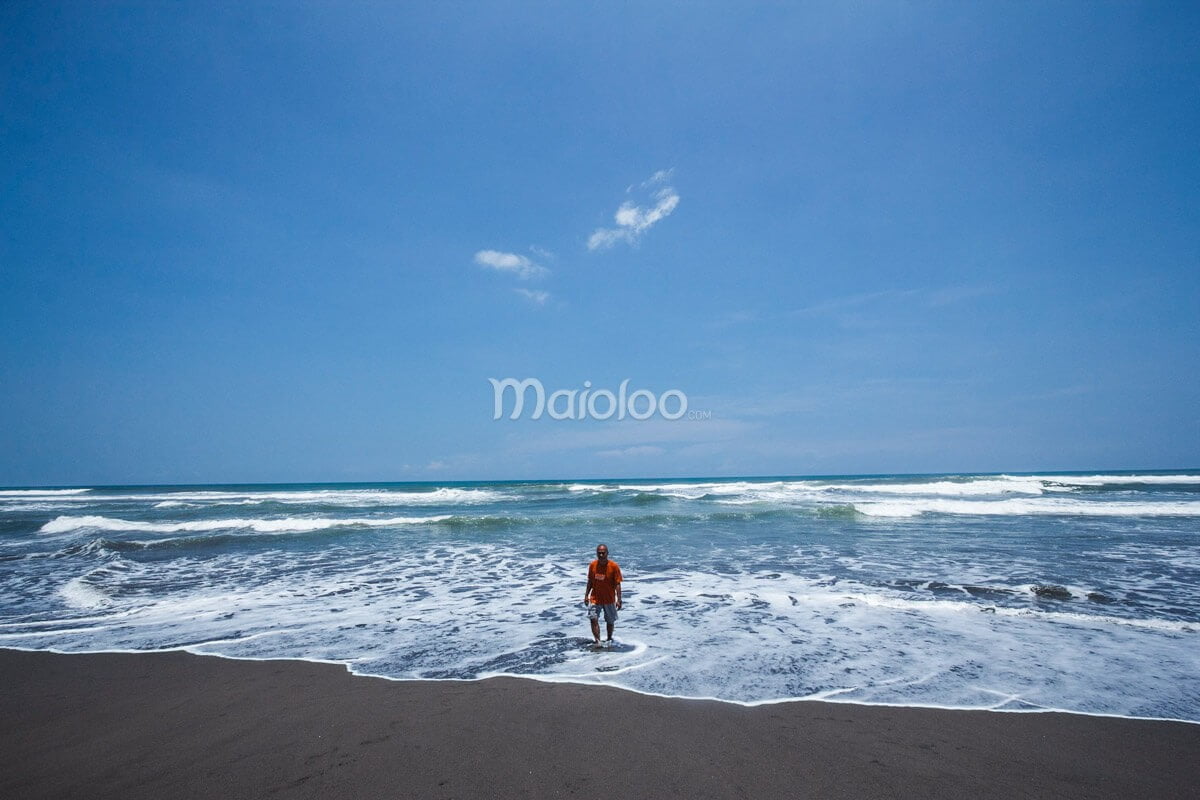 A man standing at the edge of the water on Parangkusumo Beach, with waves and a clear blue sky in the background.