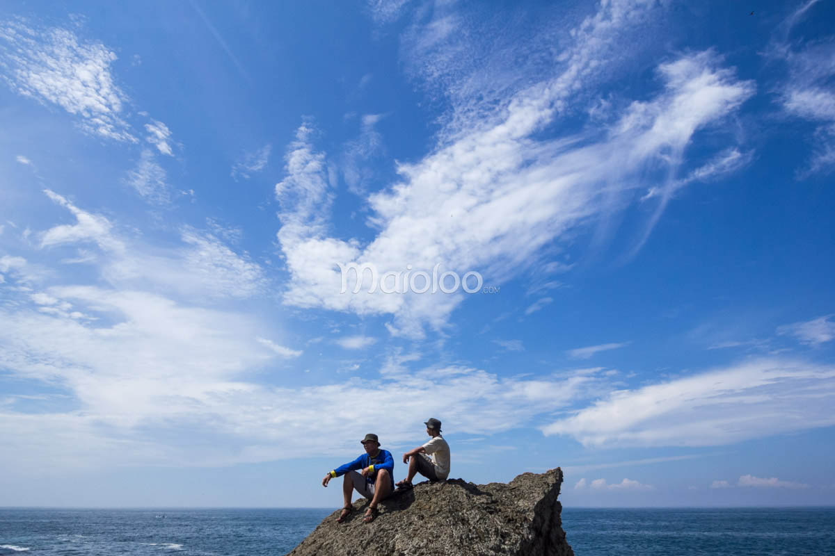 Tourists sitting on a rocky outcrop, enjoying the panoramic view of Wohkudu Beach under a bright blue sky.