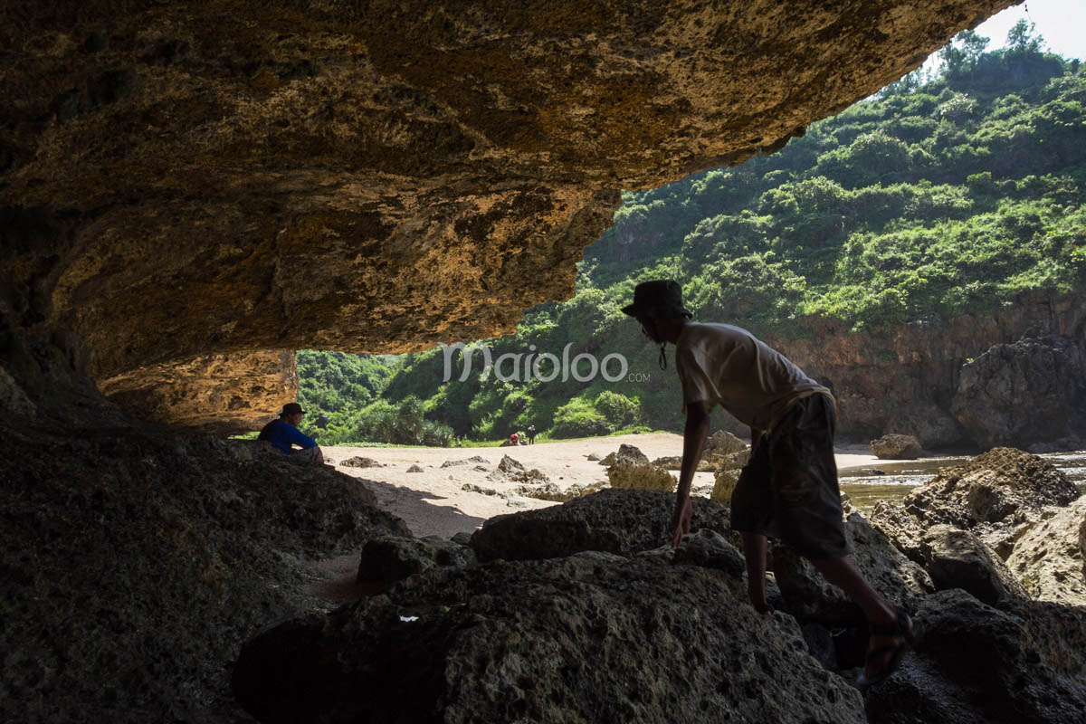 Tourists exploring the rocky area under a cliff at Wohkudu Beach with green hills in the background.