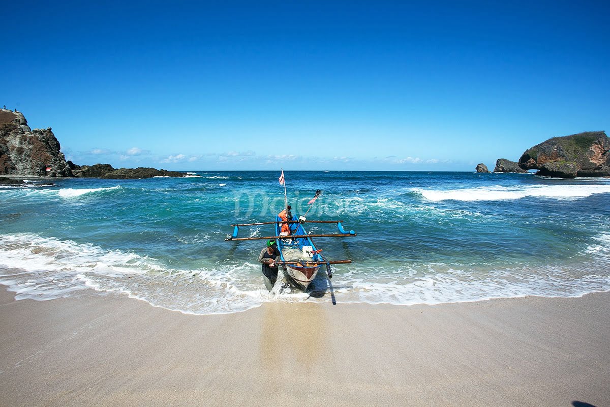 Fishermen bringing their boat ashore at Siung Beach with the ocean and rocky cliffs in the background.