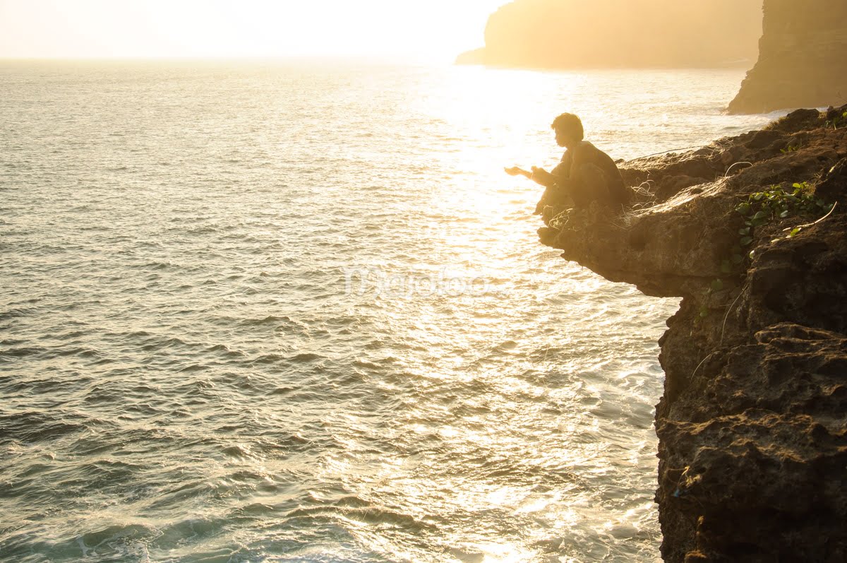 A man fishing on the edge of a cliff at Timang Beach during sunset.