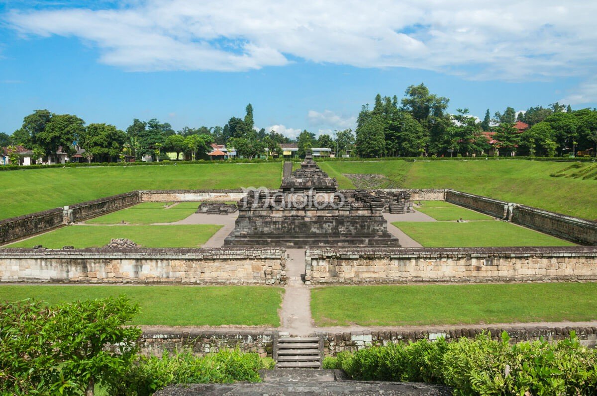 A distant view of the main structure of Sambisari Temple, surrounded by green grass and low stone walls.