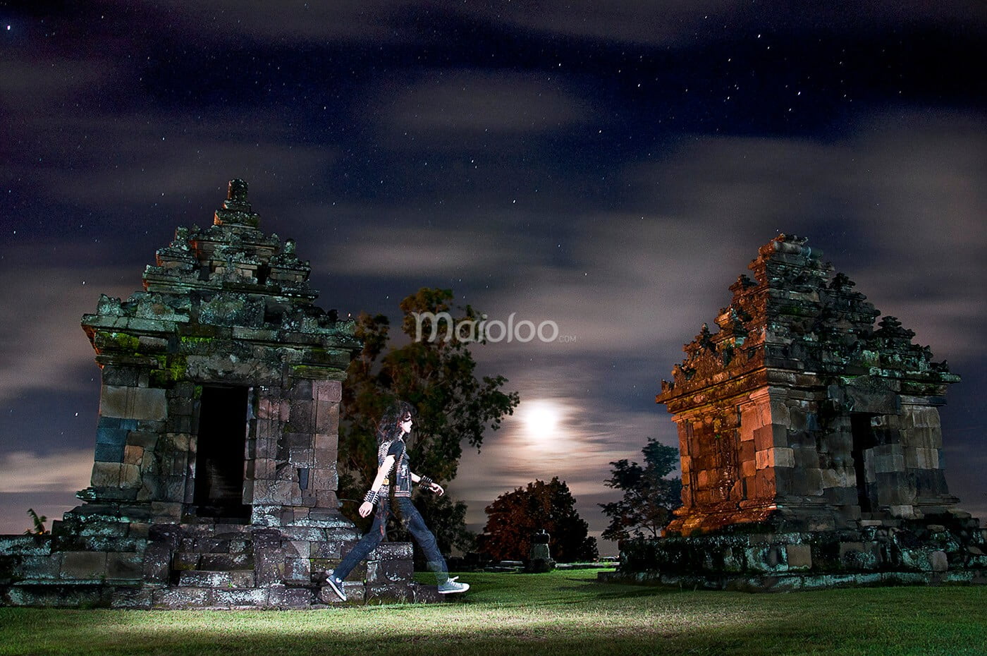 A person walking at night between ancient temple structures in Ijo Temple, with a bright moon and starry sky.