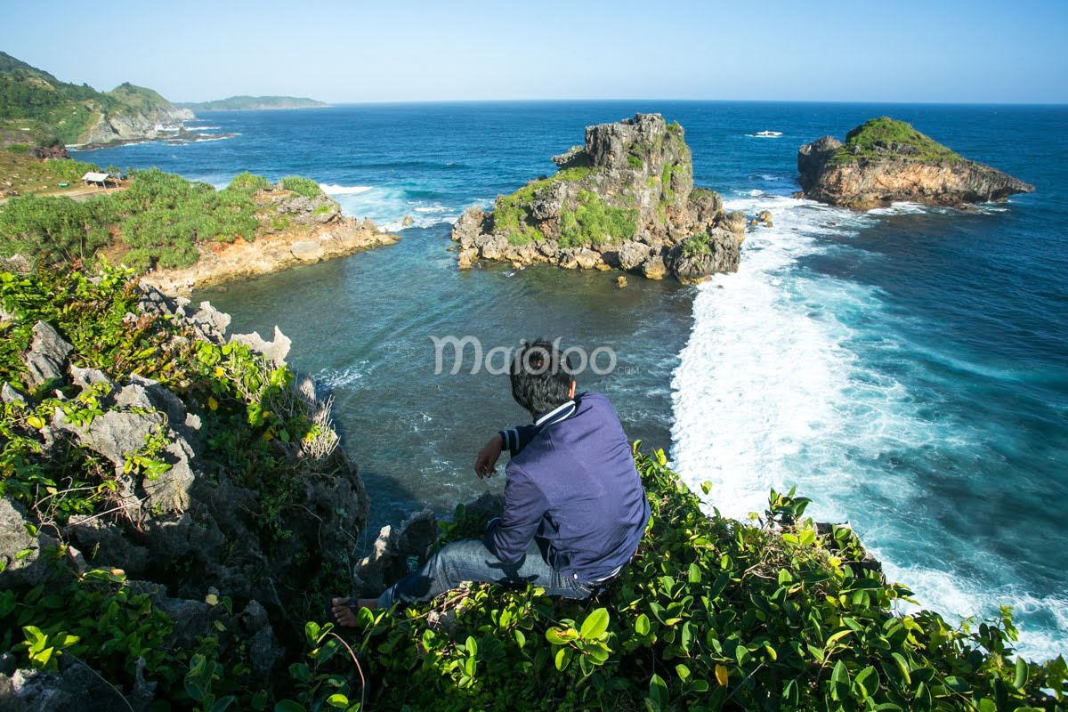 A person sitting on a cliff overlooking Nglambor Beach with clear blue waters and rocky formations.