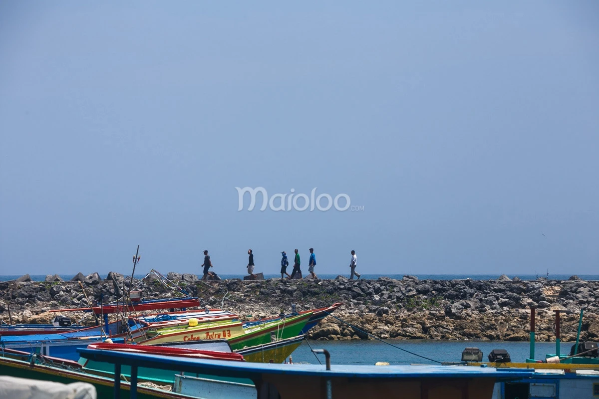 People walking along a rocky breakwater with colorful fishing boats in the foreground at Sadeng Beach.