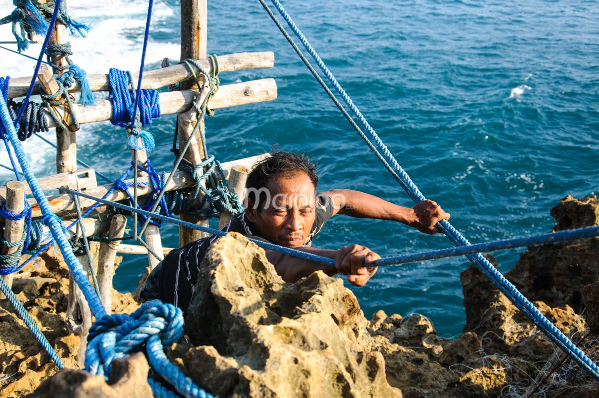 A man pulling ropes to move a gondola at the edge of a cliff with the ocean in the background.