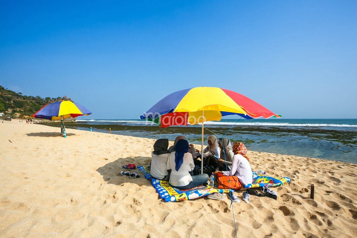 Visitors at Pok Tunggal Beach sitting under colorful umbrellas on the white sand.