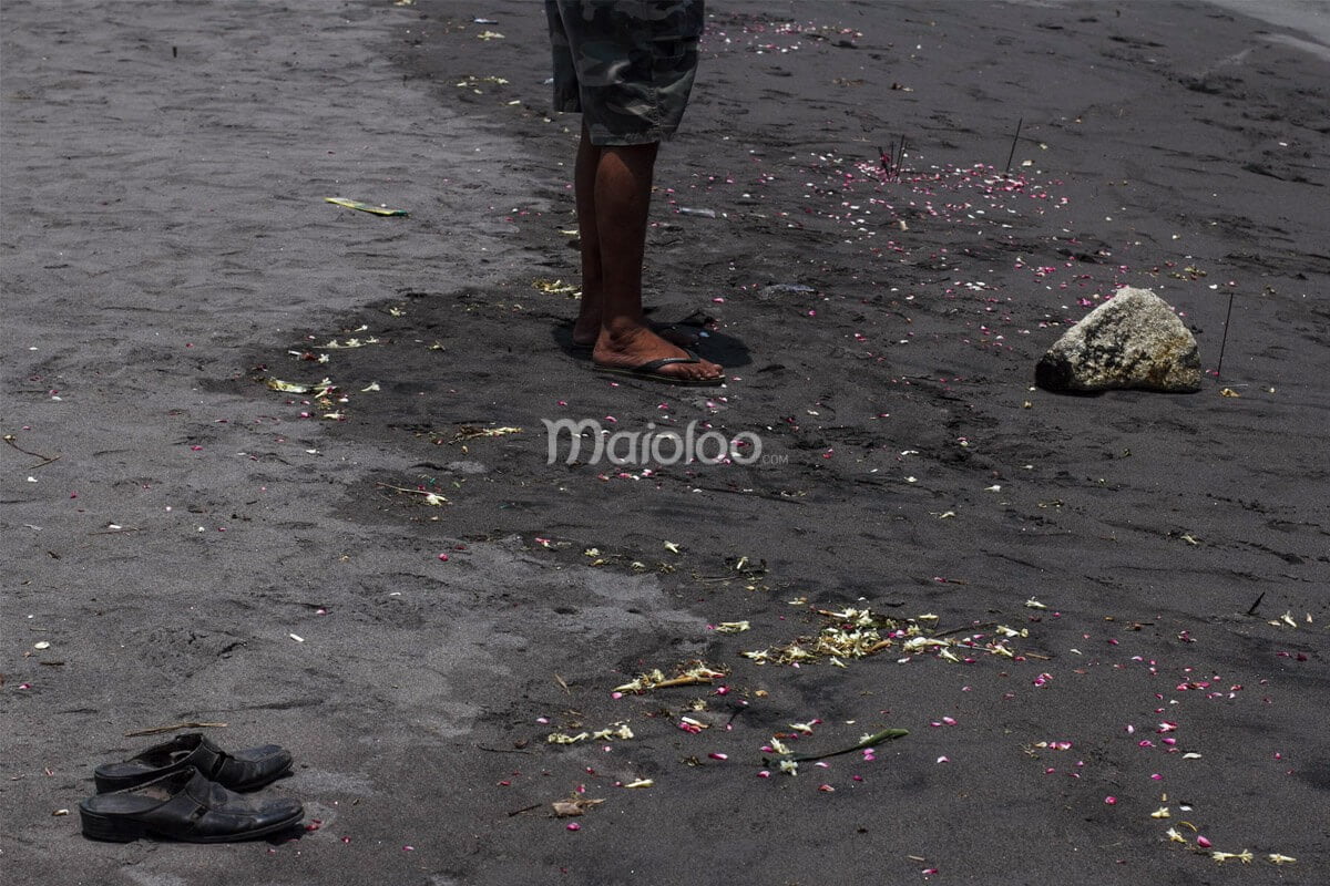 A person standing barefoot on Parangkusumo Beach, surrounded by flower petals and incense sticks on the black sand.