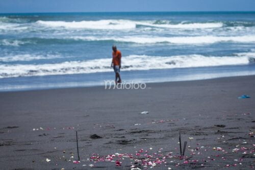 Incense sticks and flower petals on the black sand beach of Parangkusumo, with a man walking near the waves in the background.