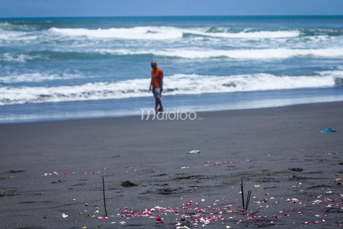 Incense sticks and flower petals on the black sand beach of Parangkusumo, with a man walking near the waves in the background.