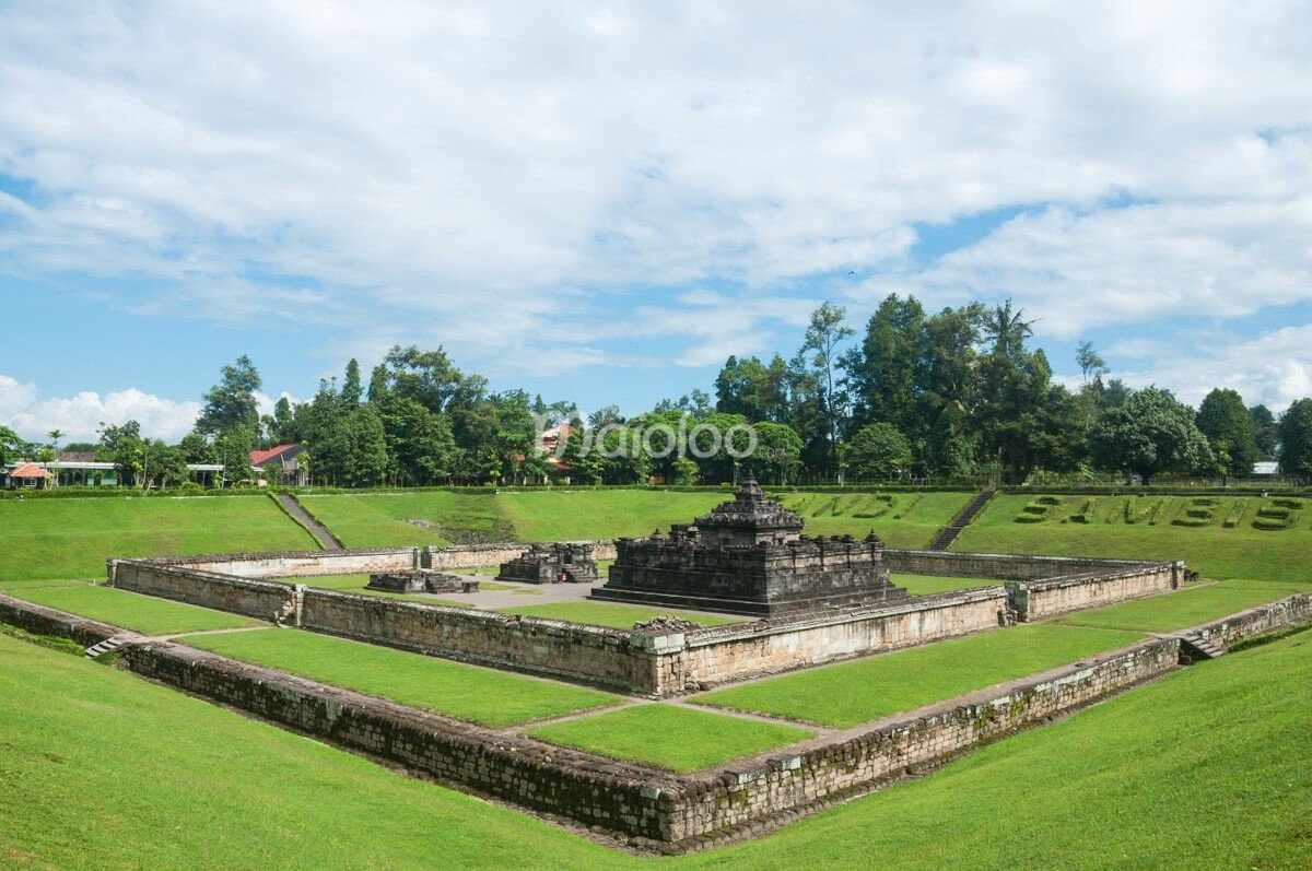Sambisari Temple, a Hindu temple submerged in a green pit, under a bright sky with scattered clouds.