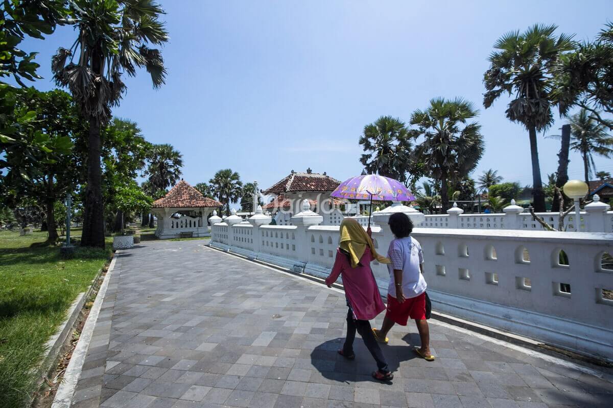 Two visitors walking with an umbrella at Cepuri Parangkusumo, surrounded by palm trees and traditional buildings.