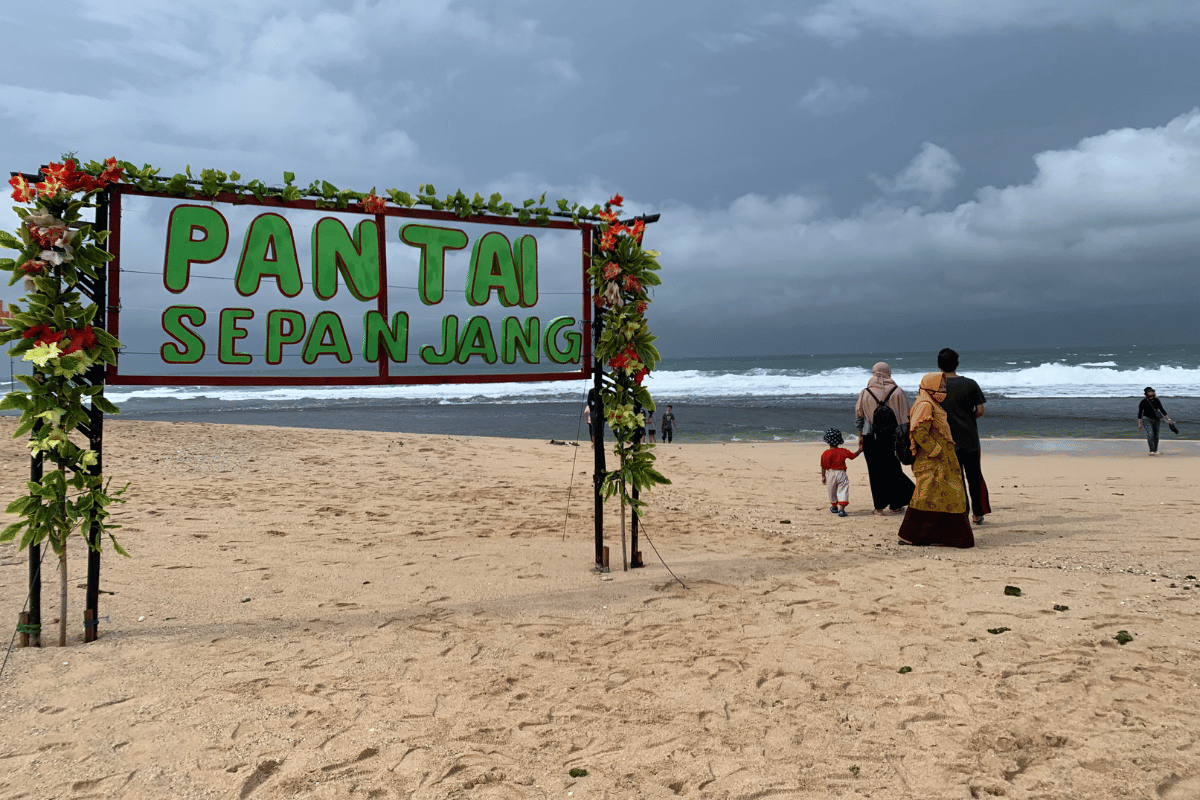 A signboard at Sepanjang Beach with visitors walking on the sandy shore.