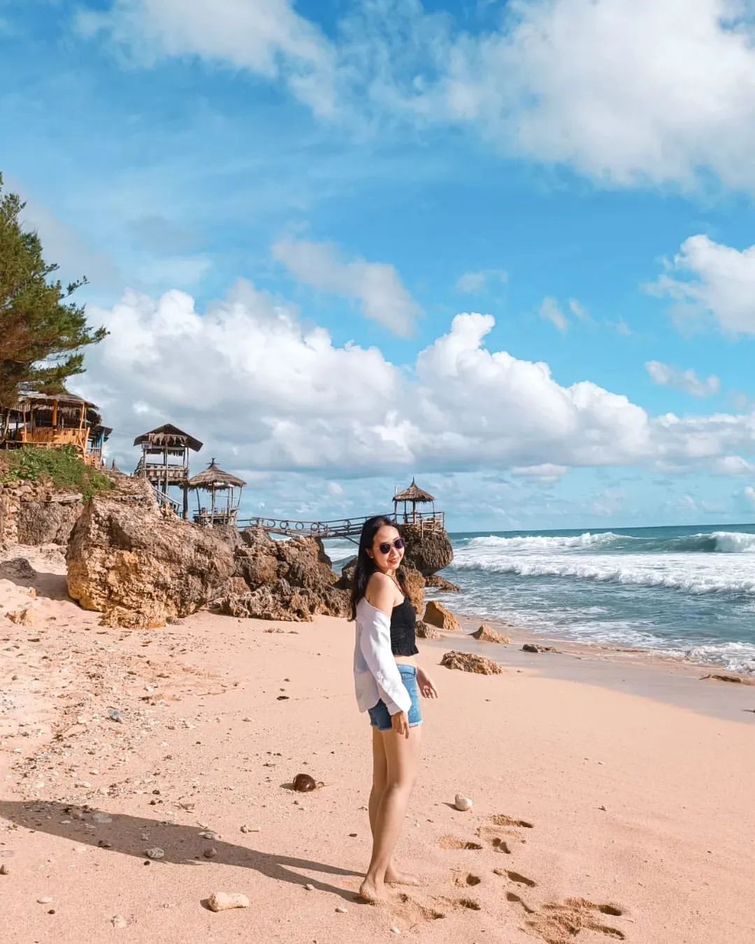 Woman standing on the sandy shore of Watulawang Beach with rocky outcrops and a bamboo bridge in the background.