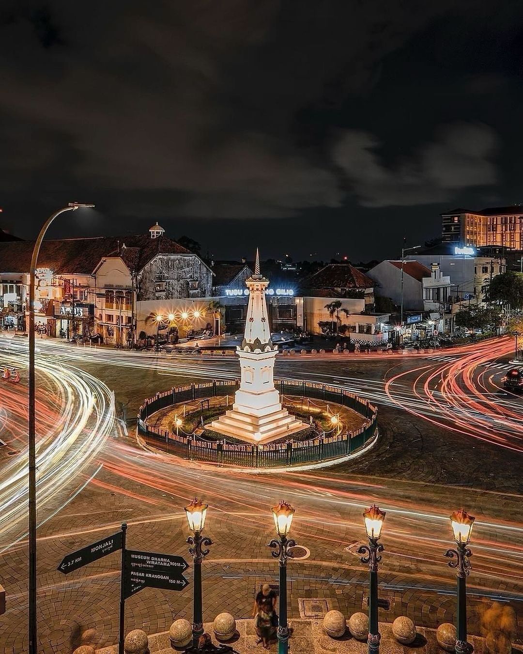 An aerial view of the Tugu Yogyakarta monument at night, surrounded by a roundabout with light trails from passing vehicles.