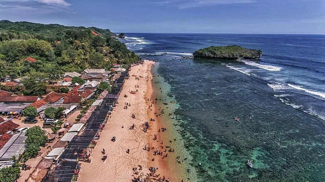 Aerial view of Sadranan Beach showing people enjoying the sandy shore and clear waters with surrounding green hills and a small island.