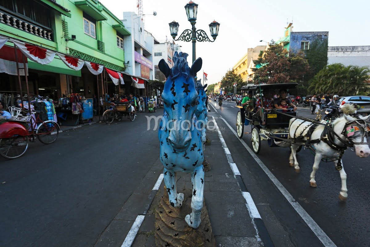 A blue horse sculpture on the sidewalk of Malioboro Street with horse-drawn carriages and shops in the background.