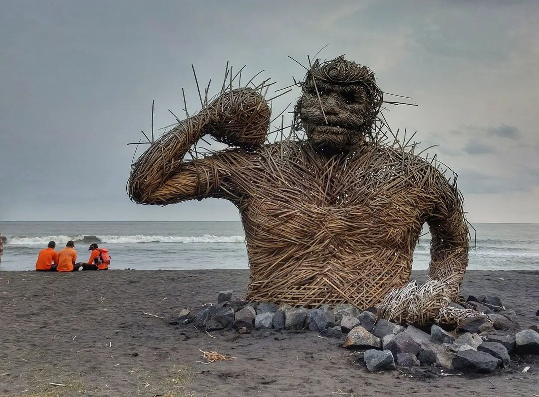 A large bamboo gorilla sculpture on Trisik Beach with people sitting in the background.