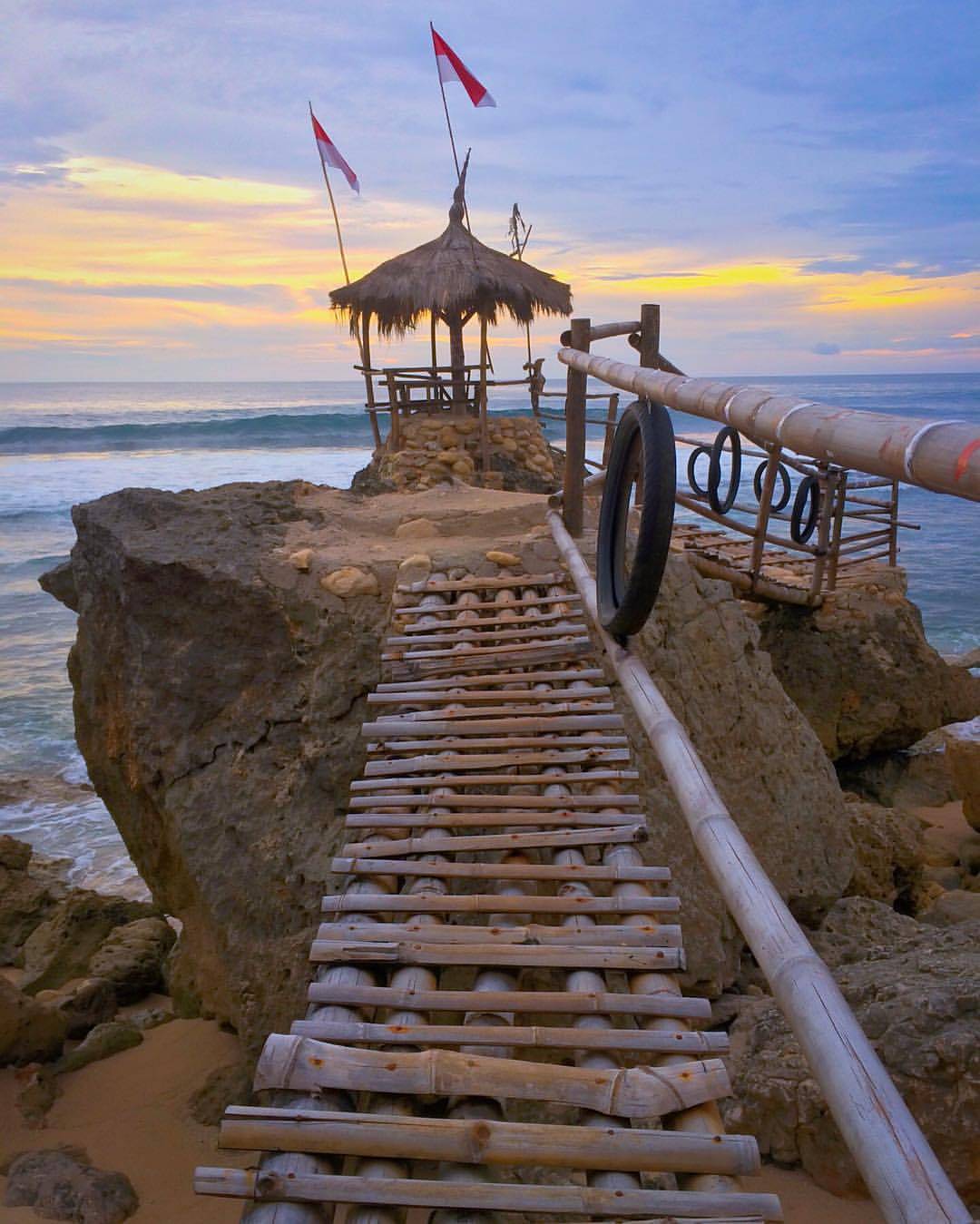 Bamboo bridge leading to a small hut on rocks at Watulawang Beach during sunset.