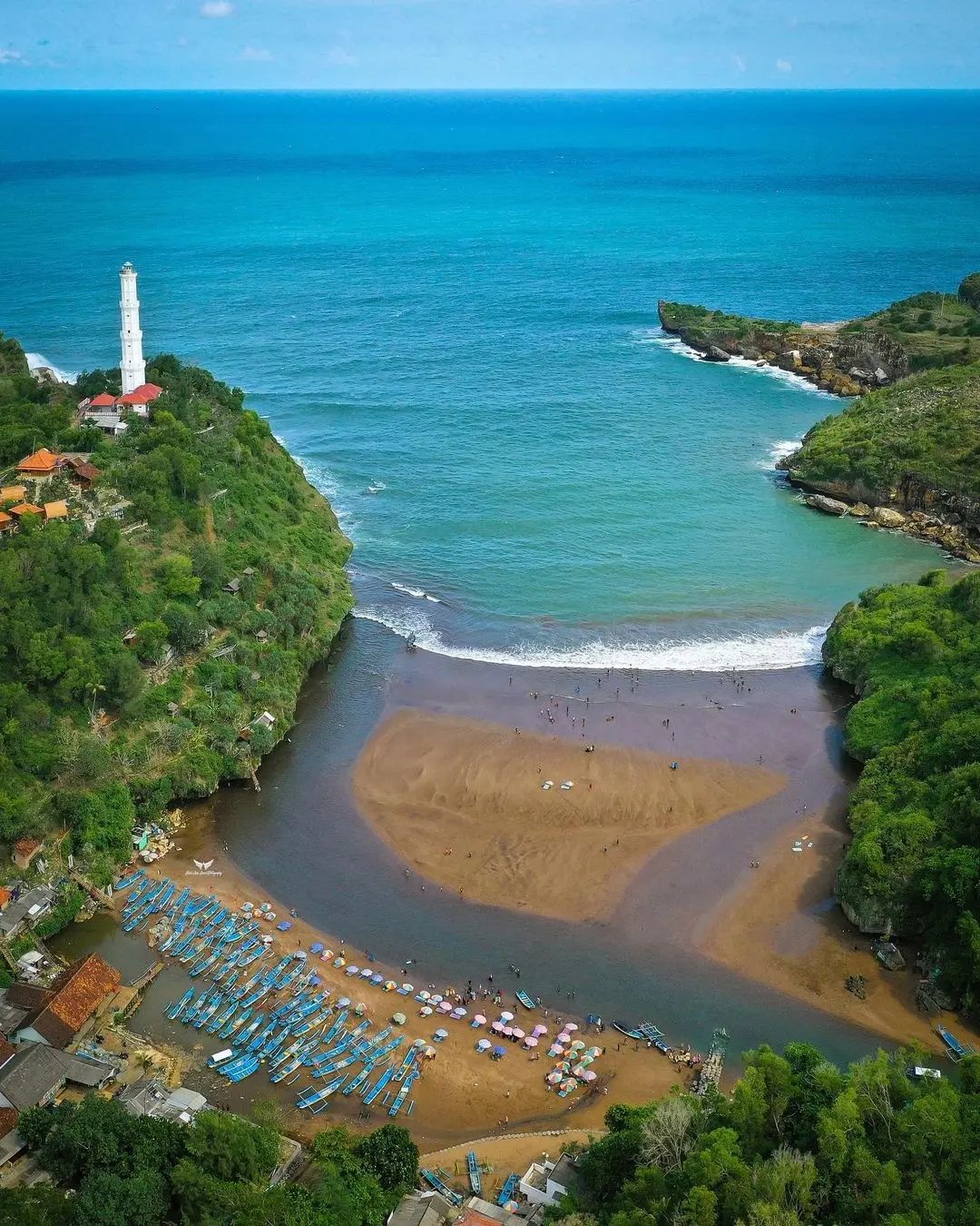 Aerial view of Baron Beach with a lighthouse on the hill, turquoise sea, and fishing boats on the shore.