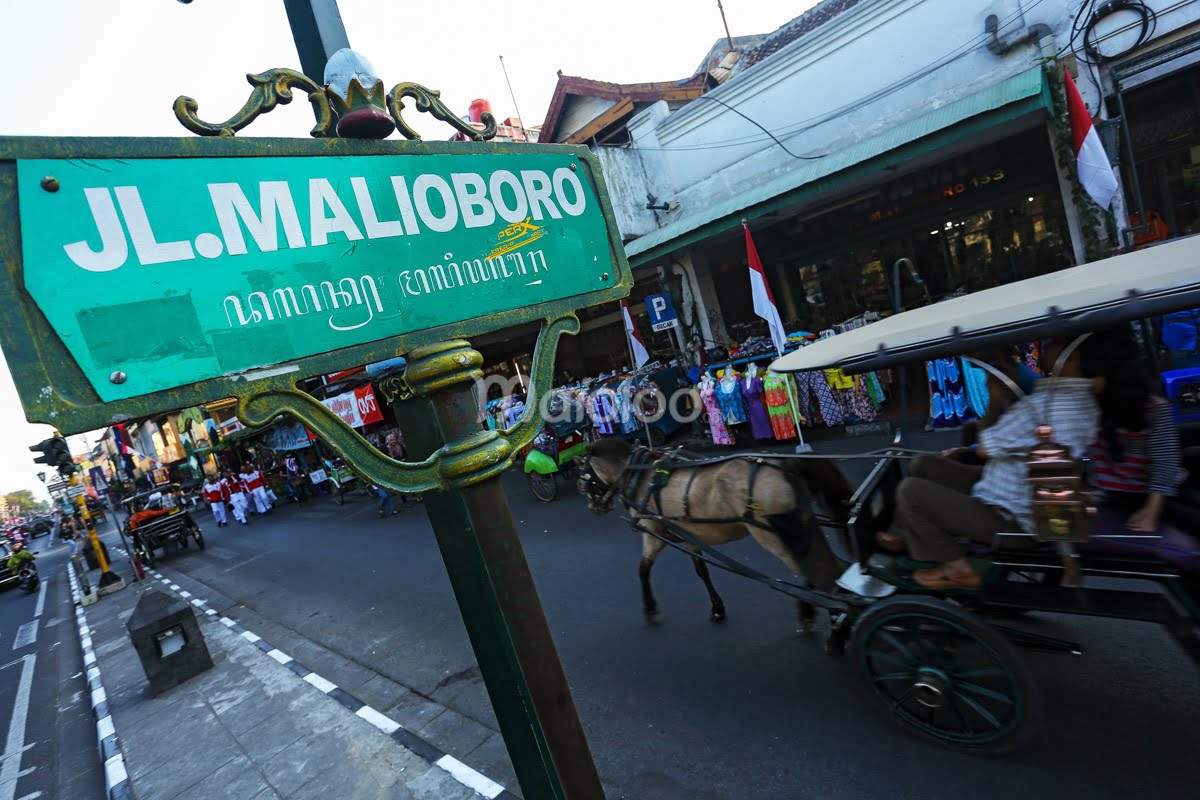 A green street sign that reads "Jl. Malioboro" with a horse carriage and shops in the background.