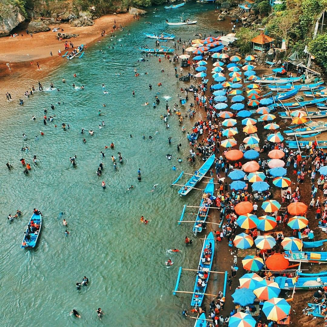 Crowded Baron Beach with colorful umbrellas, fishing boats, and people swimming in the turquoise water.