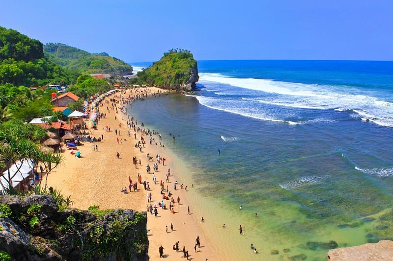 Aerial view of Indrayanti Beach with visitors enjoying the water and sandy shoreline, surrounded by lush greenery.