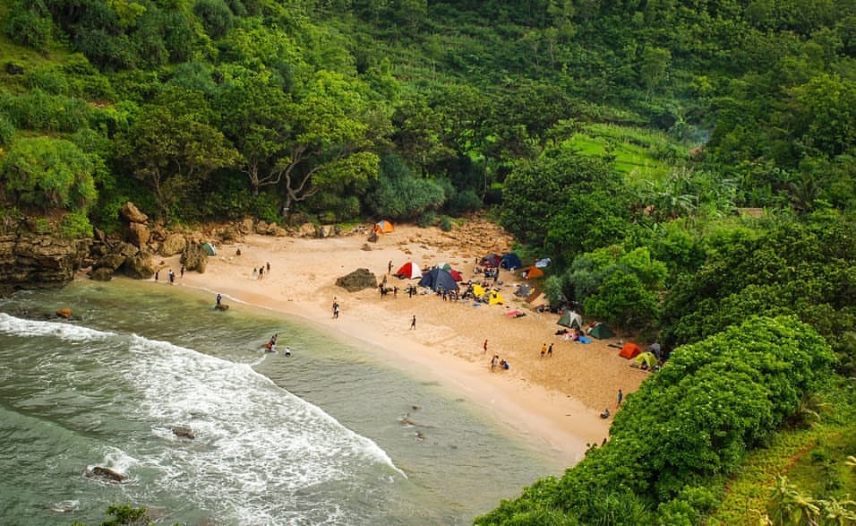 Aerial view of Ngetun Beach with several tents and campers on the sandy shore surrounded by lush greenery.