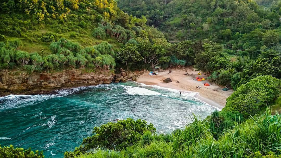 Aerial view of Ngetun Beach surrounded by green hills with tents set up on the sandy shore.