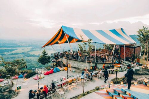 A vibrant outdoor restaurant with colorful seating and a striped tent at Obelix Hills, overlooking lush green landscapes.