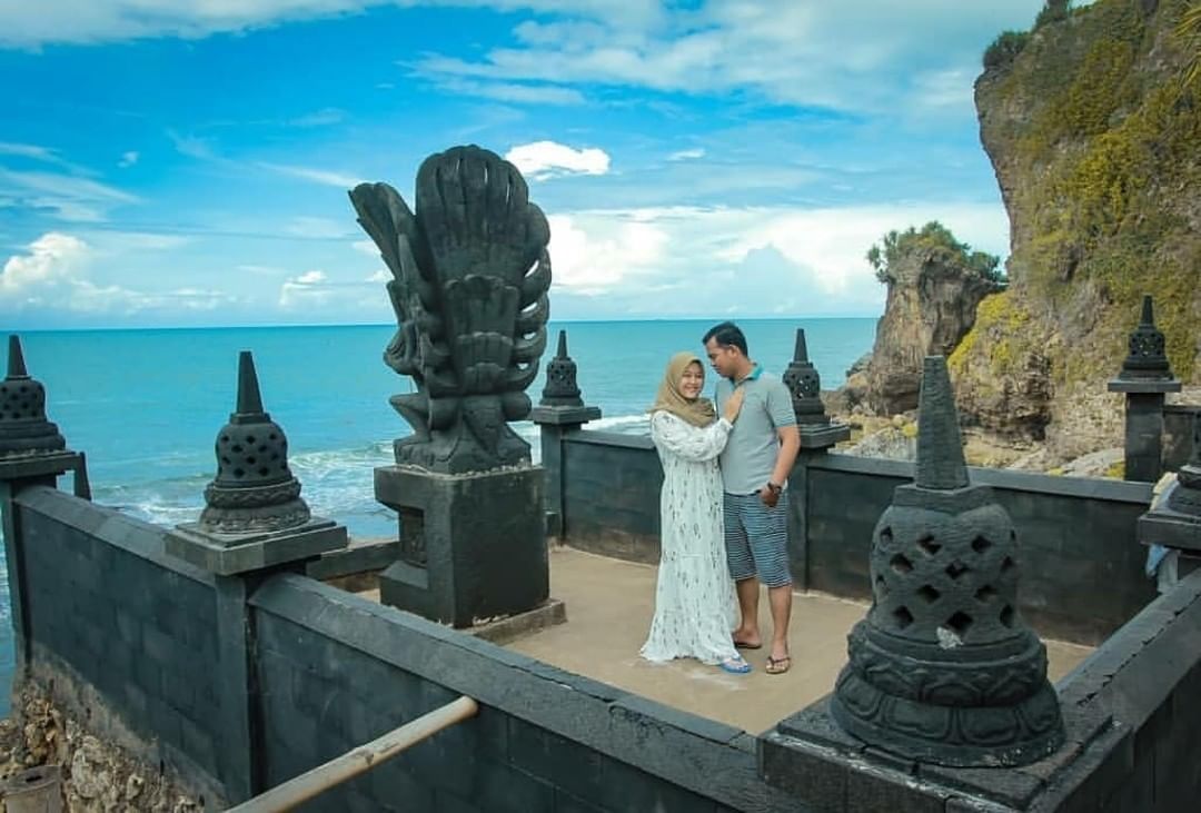 A couple posing near a Hindu statue on a cliff at Ngobaran Beach with the ocean in the background.