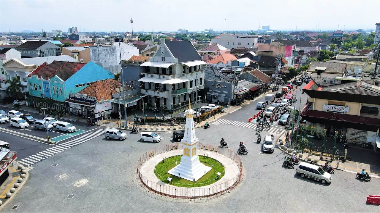 An aerial view of the Tugu Yogyakarta monument during the day, situated in the center of a busy roundabout surrounded by buildings and traffic.