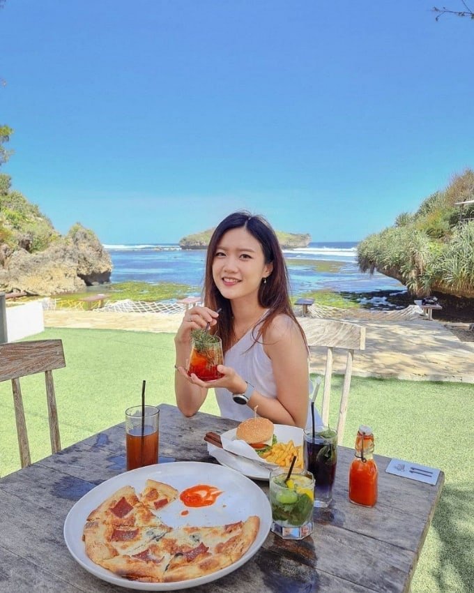 A woman enjoying a meal and drink at an outdoor table with a scenic ocean view at Slili Beach.