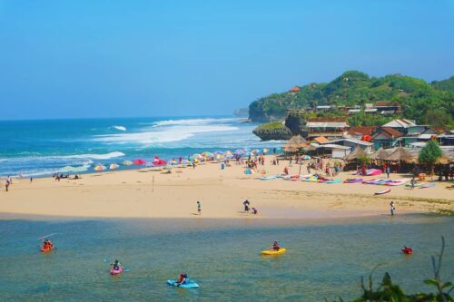 A sunny beach with colorful umbrellas, people kayaking, and enjoying the sand and sea.