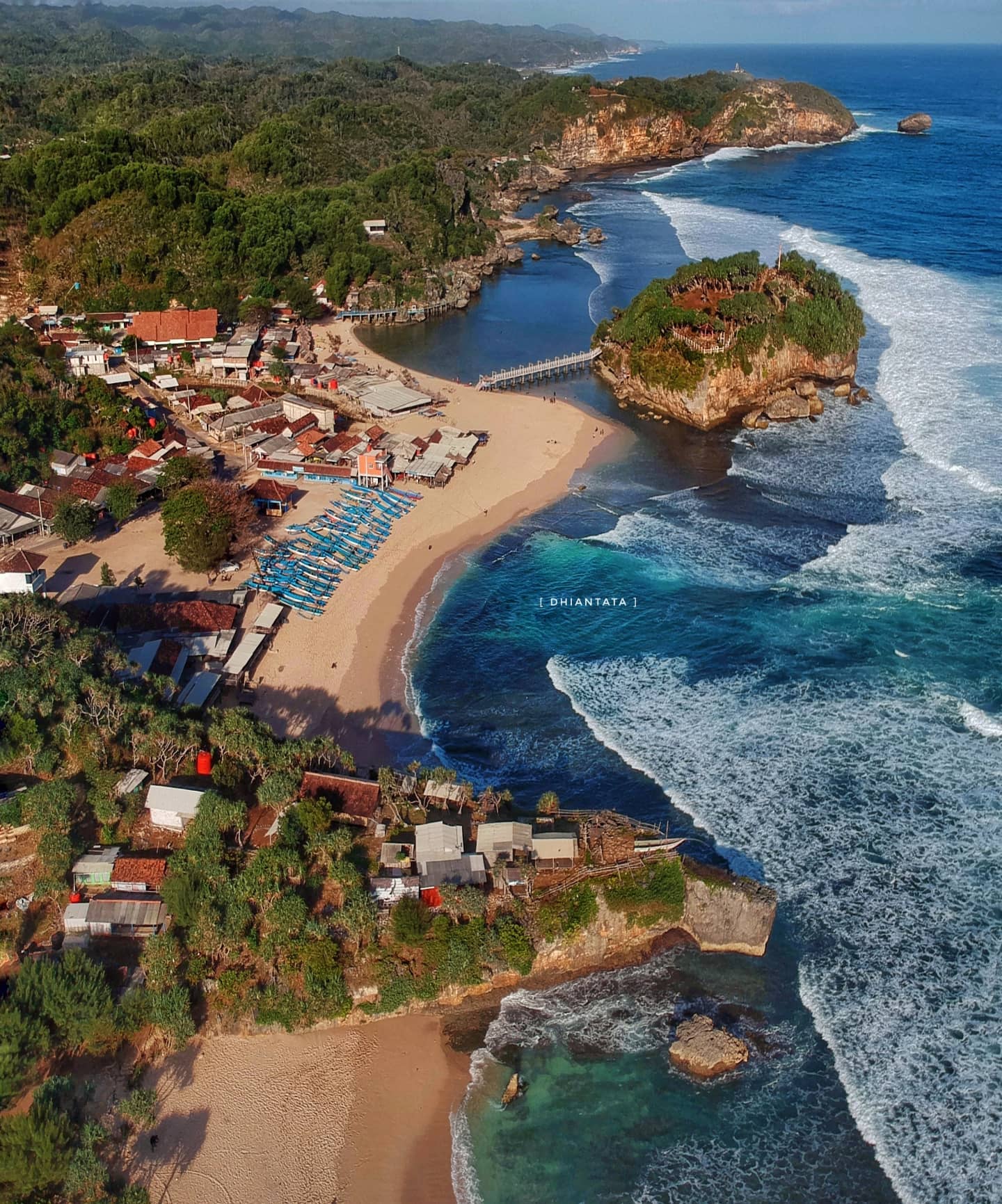 An aerial view of a beach with a small island, clear water, and surrounding green hills.