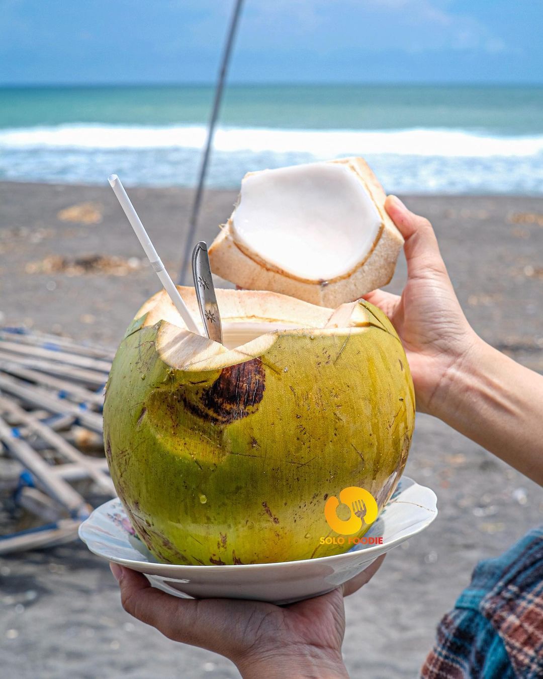 Person holding a fresh coconut with a spoon and straw at Depok Beach.