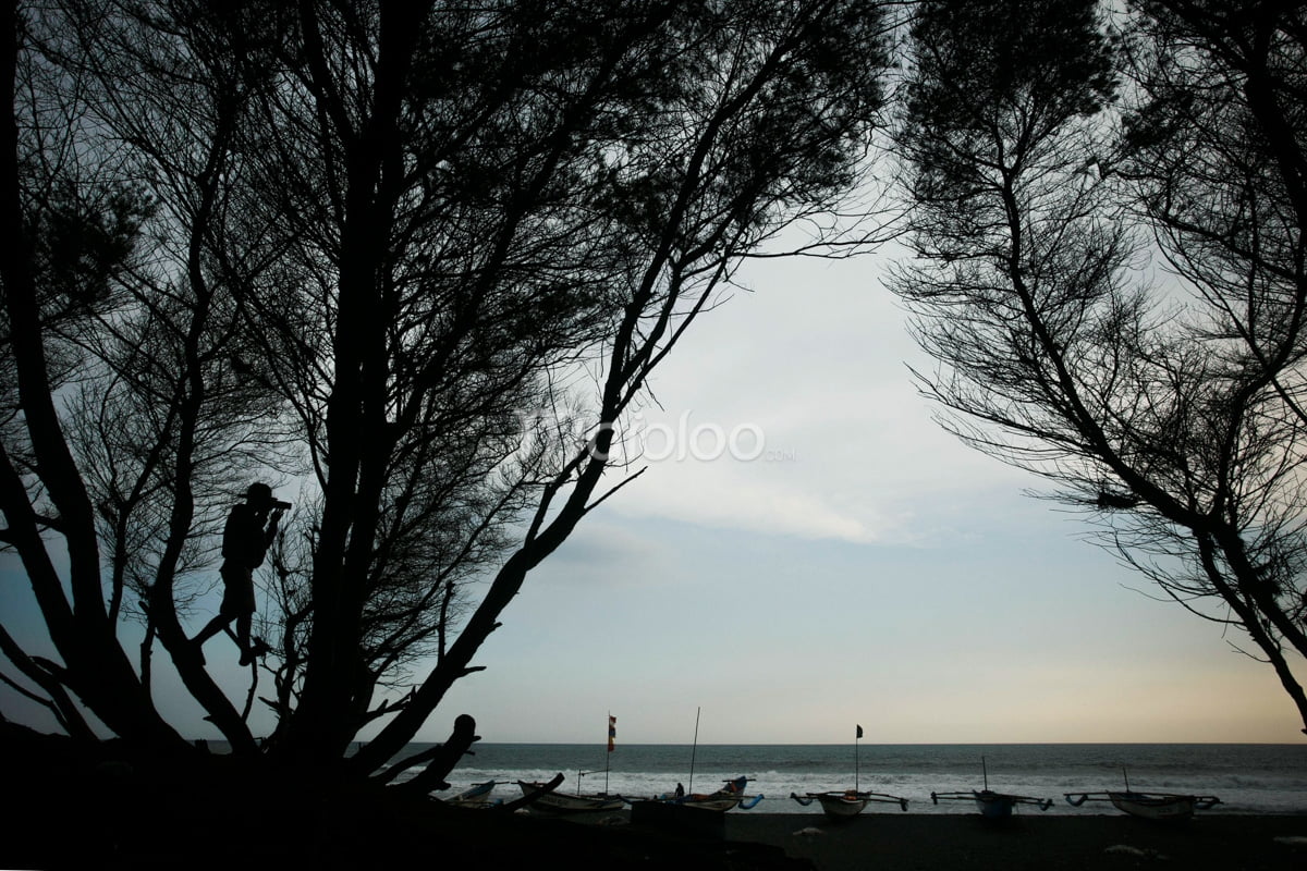 Silhouette of a person among trees at Goa Cemara Beach with boats on the shore.