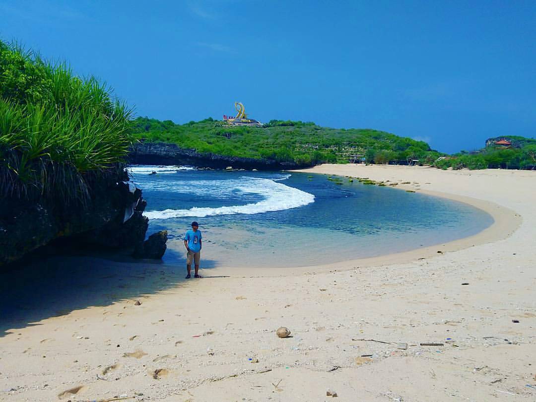 A person standing on the sandy shore of Krakal Beach with the yellow fish statue visible in the distance.