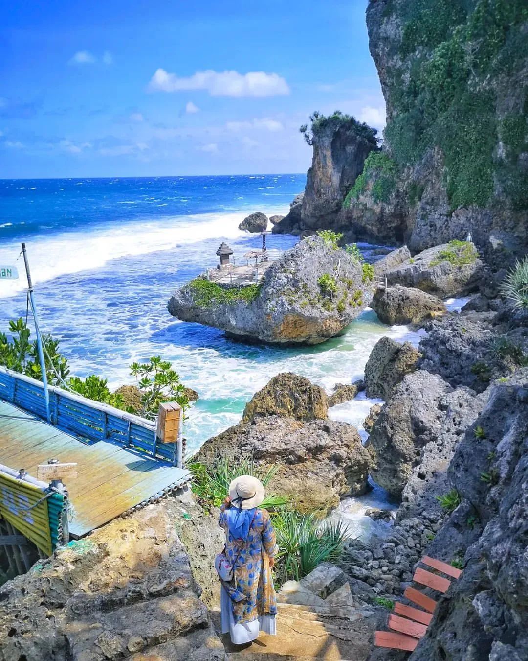 A woman in a hat and colorful dress standing on steps overlooking the rocky coastline and blue ocean at Ngobaran Beach.