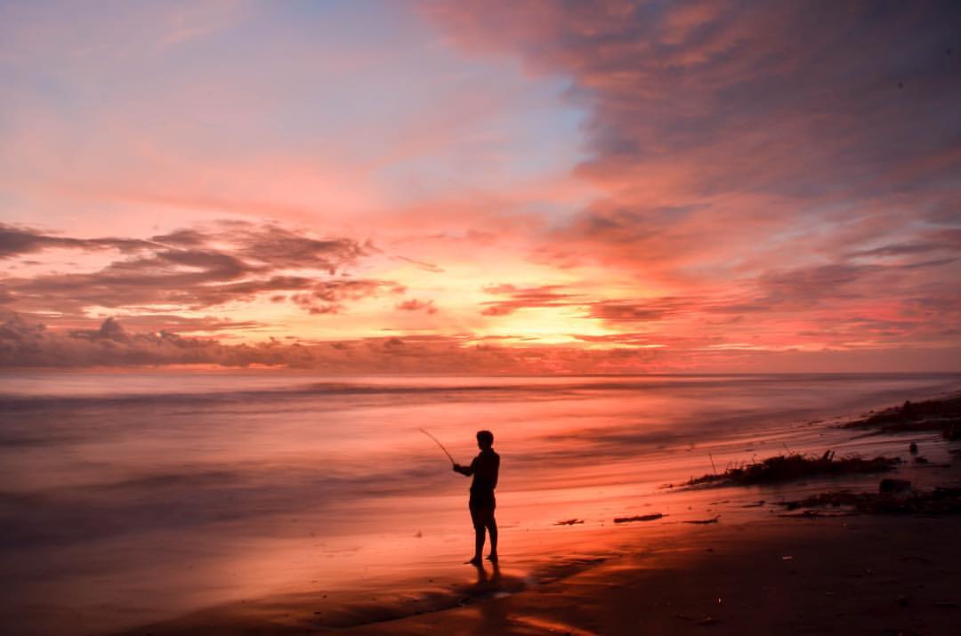 A person fishing at Depok Beach during a vibrant sunset.