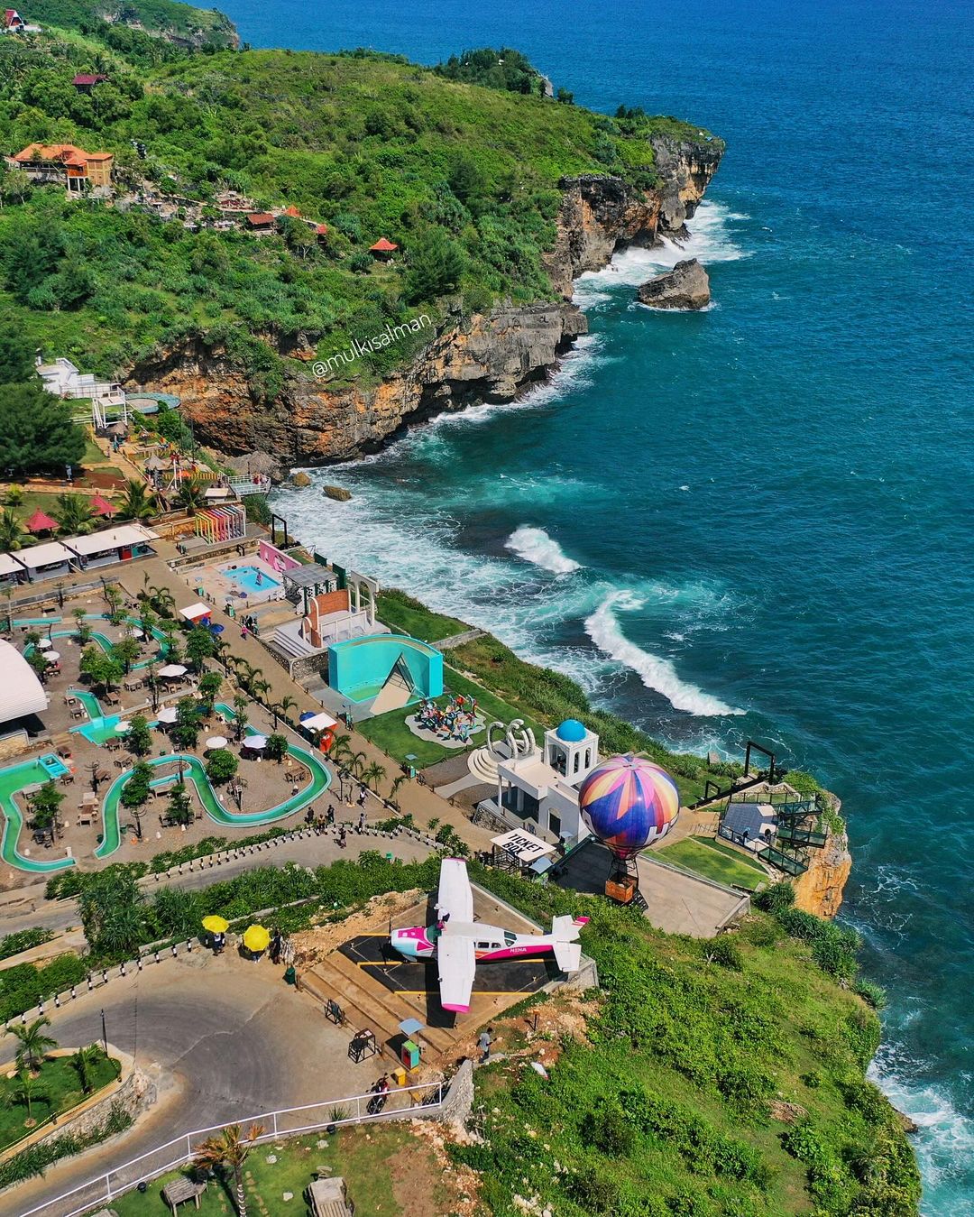 Aerial view of HeHa Ocean View with blue ocean, cliffs, and colorful attractions including a hot air balloon and small airplane set.