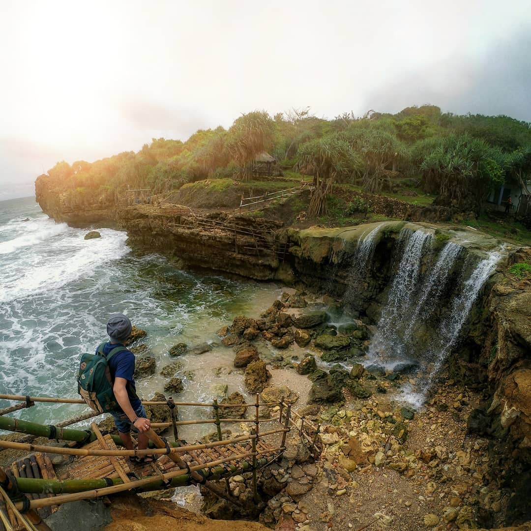 A hiker standing on a bamboo staircase, overlooking the waterfall at Jogan Beach flowing into the ocean.