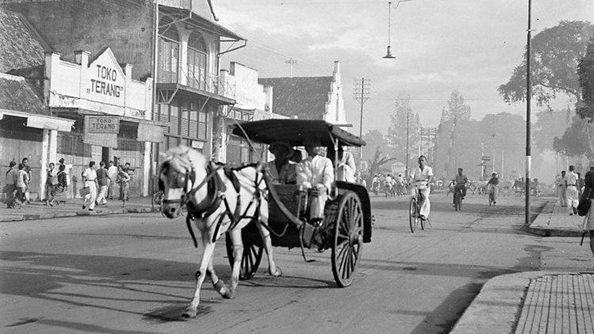 A black and white photo of a horse-drawn carriage and people on bicycles on an old street with storefronts in the background.