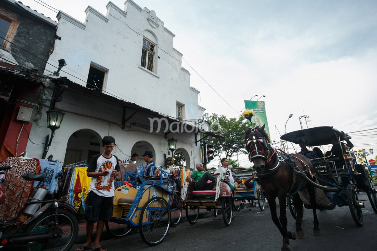 A horse-drawn carriage passing by a historical building on Malioboro Street with people and rickshaws around.