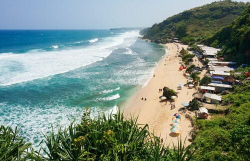 Aerial view of Indrayanti Beach with clear blue water, white sand, and colorful umbrellas along the shore.